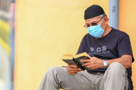Kedah/Malaysia-September 9 2020: An old man waering protective mask is seen reading english novel on the footpath near the school at alor setar kedah.のeditorial素材