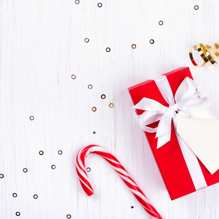 Christmas gift wrapped in red paper tied with white ribbon and with empty tag on white wooden background decorated with sparkles and candy cane lollipop. Flat lay, top view.の写真素材