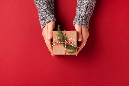Females hands in pullover holding Christmas gift box decorated with evergreen branch on red background. Christmas and New Year concept.の写真素材