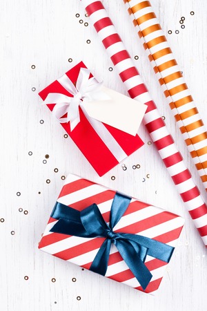 Christmas gifts wrapped in red and striped paper tied with white and blue ribbon, and tag on white wooden background decorated with sparkles. Flat lay, copy space.の写真素材
