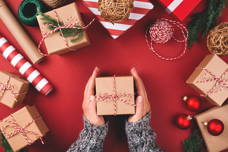 Christmas background with gift boxes, clews of rope, papers rools and decorations on red. Preparation for holidays. Top view with copy space. Womans hands holding gift box.の写真素材