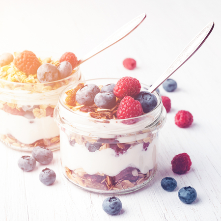 Two jars with tasty parfait made of granola, berries and yogurt on white wooden table. Shot at angle. Toned image.の写真素材