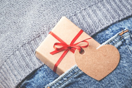 Kraft gift box with red bow and tag in shape of heart sticking out of blue jeans pocket. Valentines...