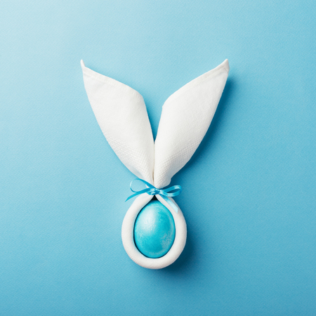 Easter bunny ears made of white napkin with egg on blue background. Minimal styled easter card concept.の写真素材