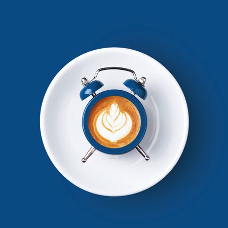 Classic alarm clock with the dial of cappuccino on blue background. Minimal styled coffee time concept.の写真素材
