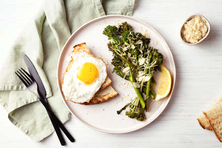 Roasted broccolini, lemon, fried egg with tost on the plate on the table decorated with napkin. Top view on healthy food.の写真素材