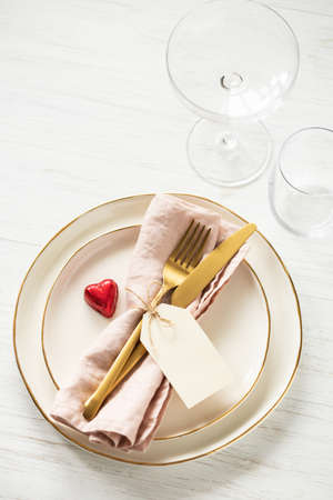 Romantic table setting with golden rim plates, candy heart, glasses and golden cutlery with tag on white table.の写真素材