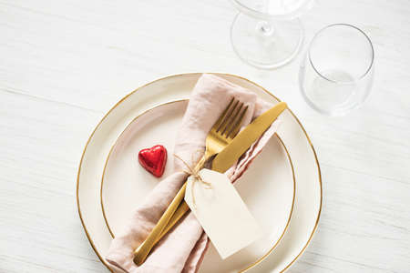 Romantic table setting with golden rim plates, candy heart, glasses and golden cutlery with tag on white table. Top view.の写真素材