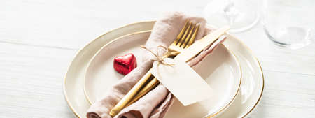Banner with romantic table setting with golden rim plates, candy heart and golden cutlery with tag on white table.の写真素材