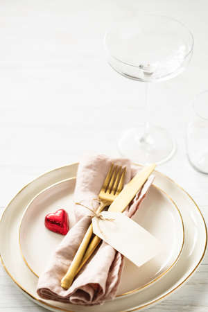 Valentine table setting with golden rim plates, candy heart, glasses and golden cutlery with tag on white table.の写真素材