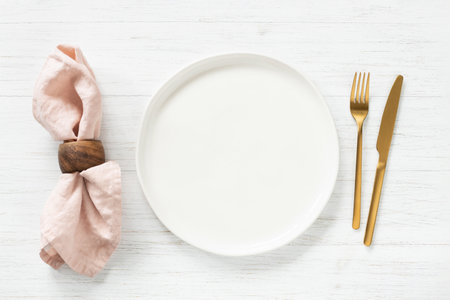 White empty plate and golden cutlery with pink napkin on white wooden table. Top view on table setting.の写真素材
