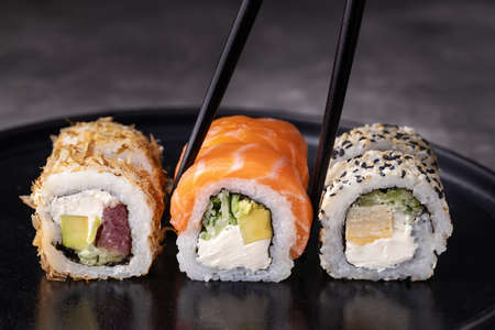 Sushi rolls set with avocado, cheese, salmon and tuna on black background. Chopsticks taking the sushi roll.の写真素材