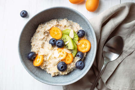 Oatmeal porridge with kumquat, blueberries, kiwi and almond flakes in a bowl. Top view on healthy breakfast on a white table.の写真素材