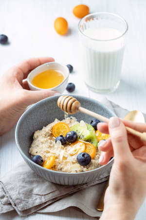 Females hands pouring honey onoatmeal porridge with blueberries, kumquat, kiwi and almond flakes.の写真素材