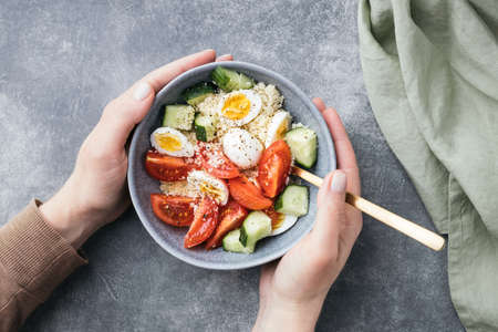 Females hands holding vegan cous cous salad bowl with cucumber, cherry tomato and quail eggs on stone table.の写真素材