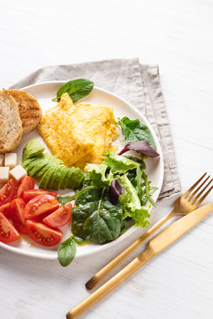 French omelet, avocado, tomatoes, salad, cheese, and bread toast on plate. Healthy breakfast on white table, angle view.の写真素材