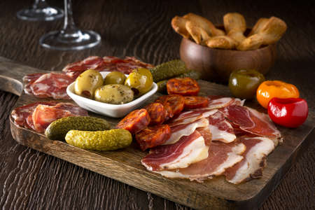 A delicious charcuterie assortment of meat, olives, gherkins, and pickled peppers with breadsticks on a wooden background.