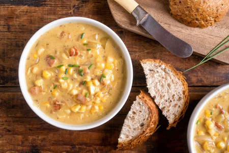 A bowl of delicious homemade bacon corn chowder with whole grain bread on a rustic wood table top.の写真素材