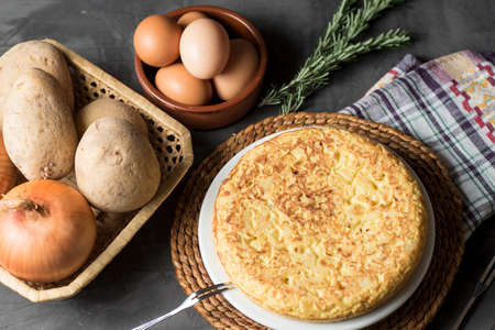 Spanish potato omelette, on a table, next to the necessary ingredients to be cooked and ready to be eatenの写真素材