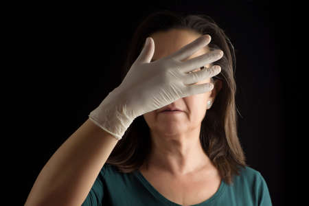 Forgive partially covering your face with one hand, with a latex glove, on a black background. Protection for contagions.の写真素材