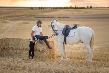 Man sitting on a straw bundle with his saddled horse and dog. Country scene.の写真素材