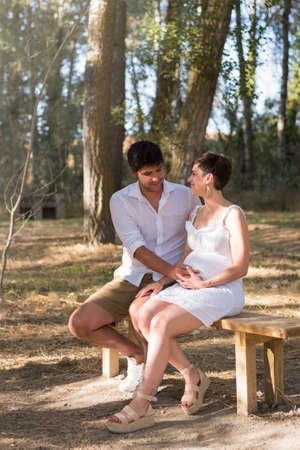 young pregnant woman, with her partner, sitting on a bench in the field.の写真素材