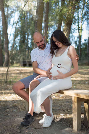 young pregnant woman, with her partner, sitting on a bench in the field.の写真素材