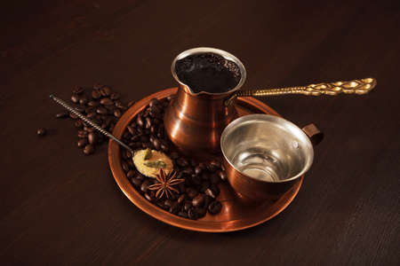 Still life, a copper coffee set consisting of a plate, a cezve, which is a turkish coffee pot, and a cup. The coffee pot is full of freshly brewed coffee. There are also coffee beans, a teaspoon of brown sugar and spices on the plate. Dark wooden backgrouの写真素材