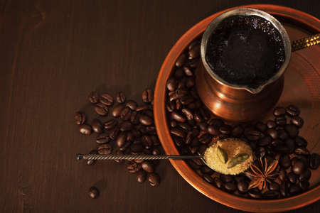 Still life, a copper coffee set consisting of a plate and a cezve, which is a turkish coffee pot. The coffee pot is full of freshly brewed coffee. There are also coffee beans, a teaspoon of brown sugar and spices on the plate. Dark wooden background.の写真素材