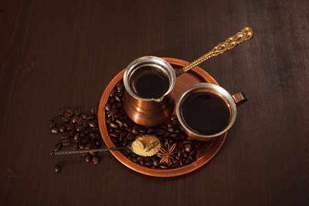 Still life, a copper coffee set consisting of a plate, a cezve, which is a turkish coffee pot, and a cup. The coffee pot is full of freshly brewed coffee. There are also coffee beans, a teaspoon of brown sugar and spices on the plate. Dark wooden backgrouの写真素材