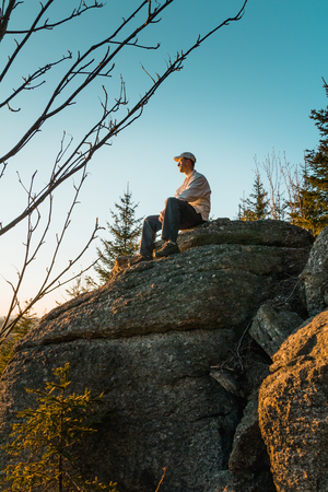 Young man hiker in white cap and shirt sit on rock from side with blue skyの写真素材