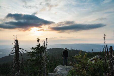 Young man tourist in white cap standing on rock at sunset in landscape, Sumava National Park and...