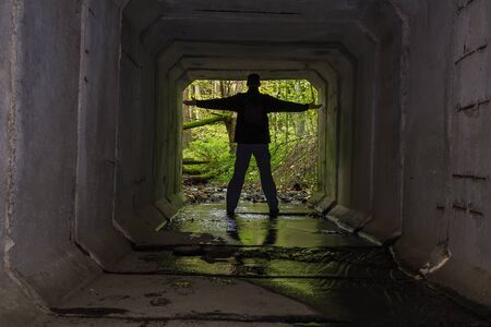 Young man standing on end of concrete stream tunnel in black jacket with backpack and open handの写真素材