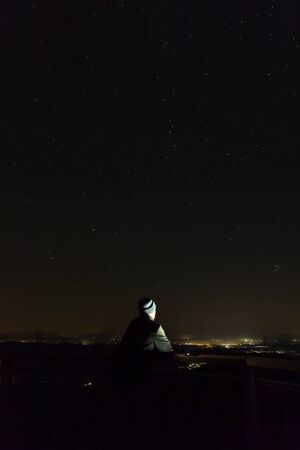 Young man looking to night sky full of stars with distant city, long exposure, Austria landscape