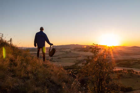 Young man standing on hill and hold backpack looking to Czech ore mountain valley at sunset landscapeの写真素材