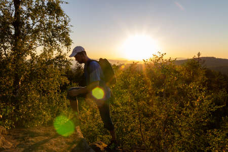 Young man with backpack hiking, walking on rock mountain in summer Czech landscape at sunset