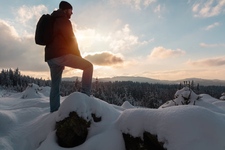 Young man from side standing on snow rock with backpack at sunset. Novohradske mountain, Czech republicの写真素材