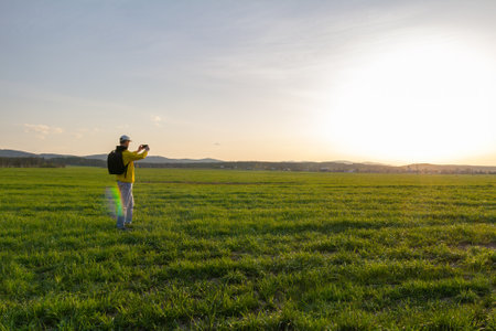 Young caucasian man in yellow jacket with backpack taking photo with mobile phone at sunset in czech spring landscapeの写真素材