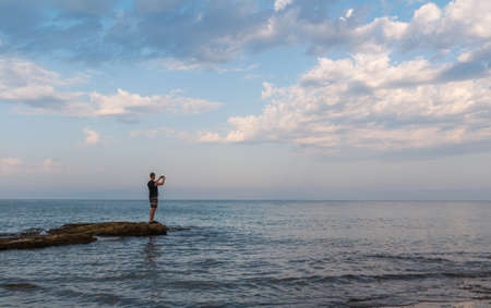 Young caucasian man in shorts taking photo with mobile phone on Croatia sea rock shore edge under cloudy sky. Side view, copy spaceの写真素材