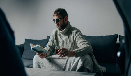 Portrait of young caucasian man in sunglasses sitting on couche reading book and drinking alcohol....