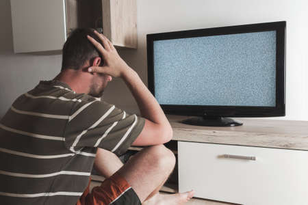 Young caucasian man sitting with head in hands looking to gray graining television. Depression, psychic, mentally ill conceptの写真素材