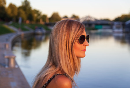 Portrait young blonde woman with sunglasses and ships in port, river Vltava, city Ceske Budejovice at sunsetの写真素材