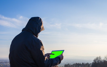Young man in jacket using tablet, pointing with finger on green screen. Landscape with distant hill at sunrise, copy space skyの写真素材