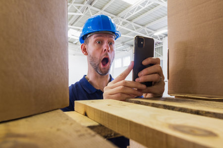 Warehouse worker in helmet shocked expression face when he sees result on phone. Stock of parcels with products, transportation conceptの写真素材