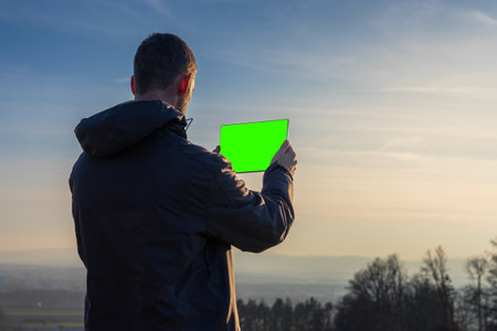 Young man in jacket using tablet with green screen. Landscape with distant hill at sunrise, copy space skyの写真素材