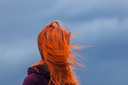 Portrait of redheaded young woman with blowing hair against blue cloudの写真素材