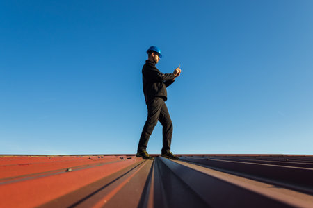 Young man worker in helmet standing on roof using mobile phone with blue sky. Roofer work backgroundの写真素材