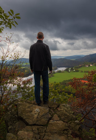Man standing on rock above river horseshoe meander under dramatic storm sky. Czech autumn landscapeの写真素材