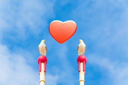 Two large seagull sitting on a wooden post. In the middle of a red heart in front of blue sky. Symbol of love.の写真素材