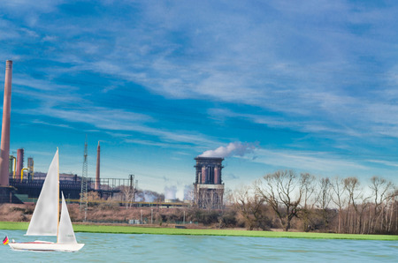 Old power plant on a river against a blue sky. On the water a sail boat. Industry, nature and leisure in harmony.の写真素材